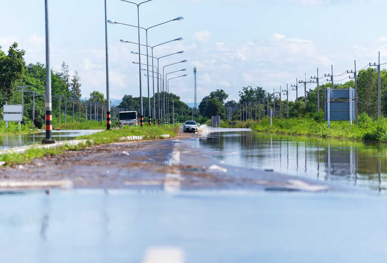 Une route partiellement inondée avec des véhicules traversant prudemment les eaux, sur une chaussée bordée de lampadaires et de verdure.